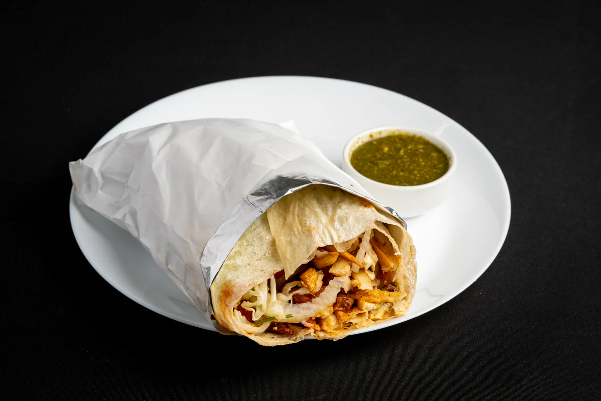 An Afghan burger rolled in paper on a white plate, open at one end to show fried potato, chicken and shredded cabbage, green chutney alongside.