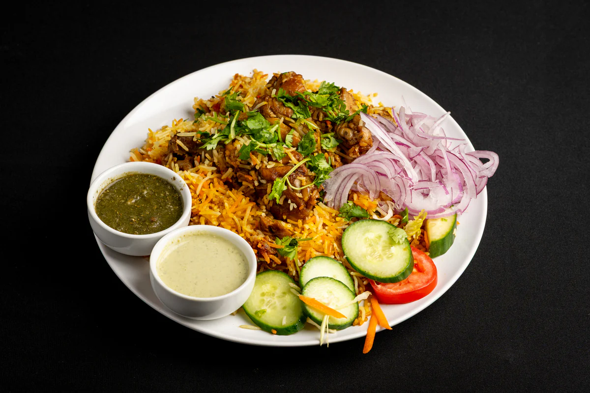 Chicken biryani on a white plate, spice-streaked rice under coriander, with sliced red onion, cucumber, tomato and two dipping sauces.