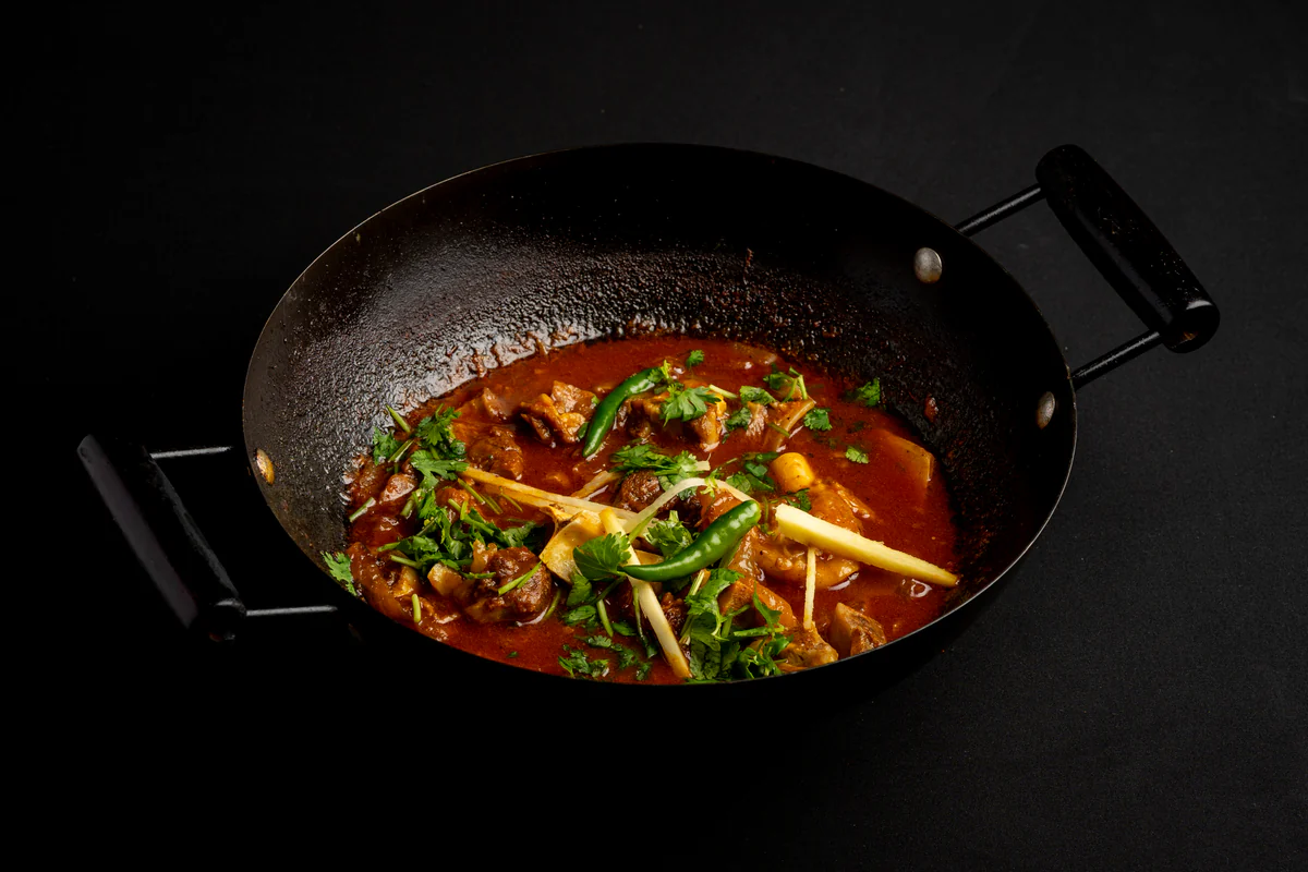Lamb curry in a black karahi, dark red sauce under coriander, julienned ginger and whole green chillies.