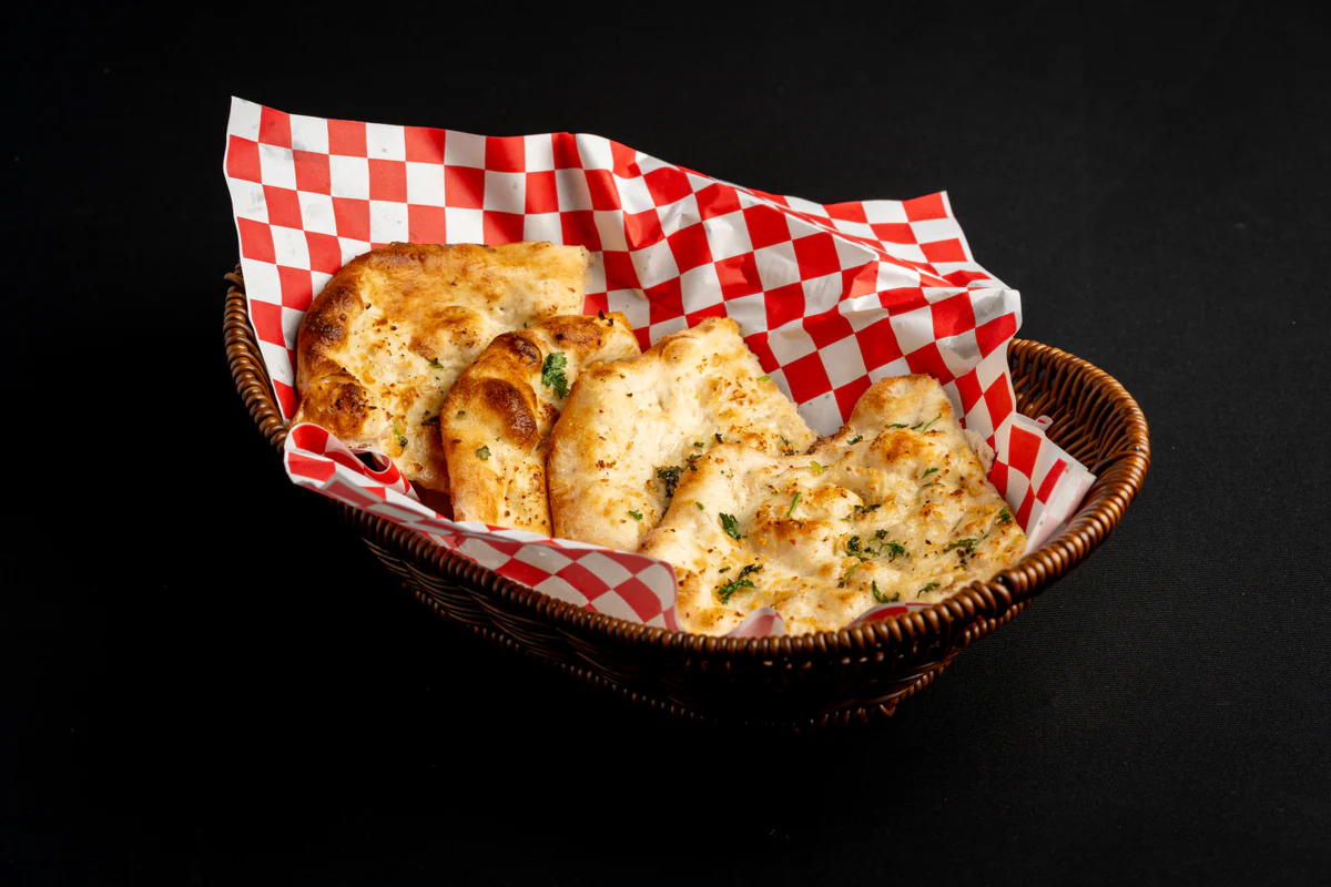 Garlic naan, herb-flecked and blistered, in a wicker basket lined with red checked paper.